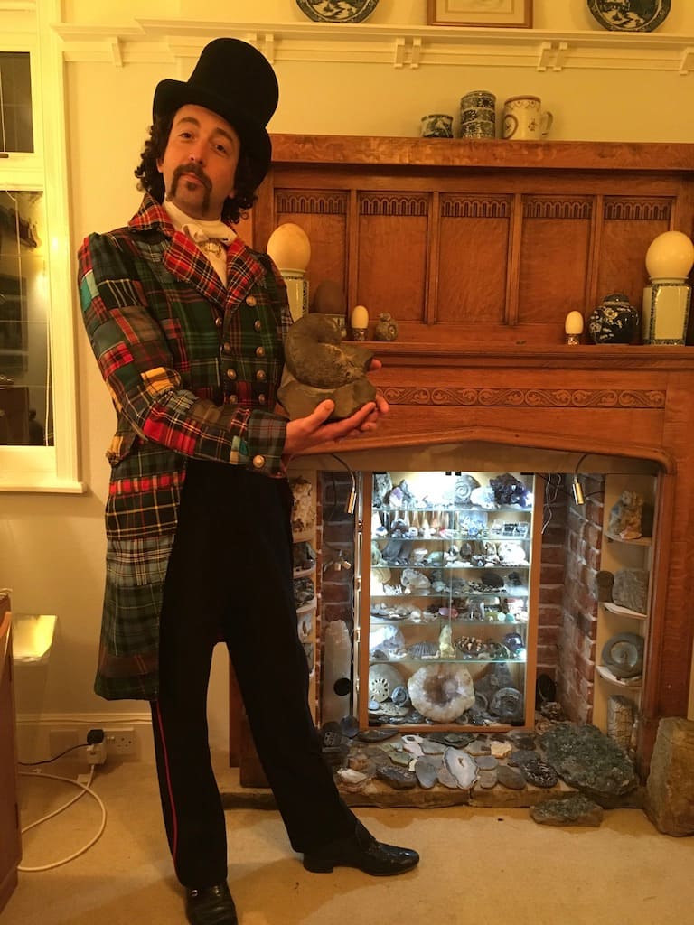 Dressed in a tartan patchwork tailcoat, The Rock Showman holds an ammonite fossil in front of a rock collection